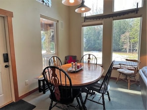 A dining area with a round wooden table, black chairs, and a bench. There are large windows providing a view of the outdoors. - Elkhorn (Packwood)
