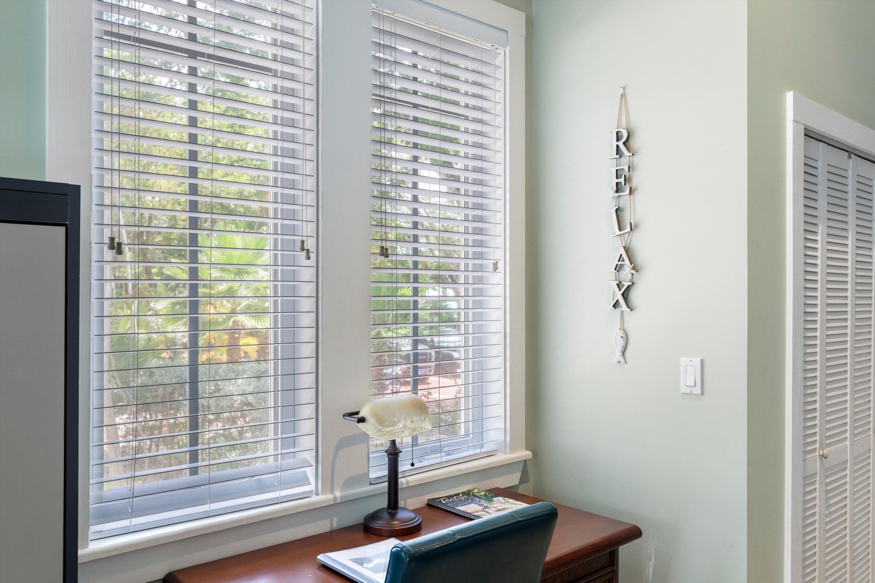 A room with two windows with blinds, a desk with a lamp, a chair, and a wall decoration.