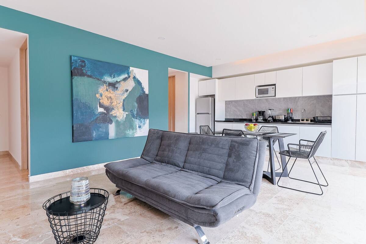Een moderne woonkamer met een grijze bank, een eettafel met stoelen en een keuken met witte kastjes en een magnetron.
