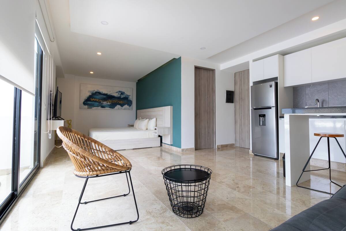 Een moderne hotelkamer met een bed, een rieten stoel, een klein zwart tafeltje en een kitchenette met een koelkast en spoelbak.