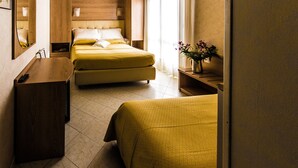 A hotel room with two beds, a wooden desk, and a small bedside table with a vase of flowers. - Hotel Baviera (Chioggia)