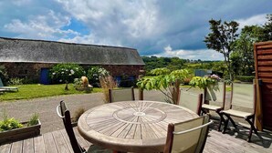 A wooden deck with a round table and chairs, a stone building in the background, and a clear sky with clouds. - Old farmhouse 100m², quiet, 500 m from the beach, sleeps 6-9 (Saint-Michel-en-Grève)