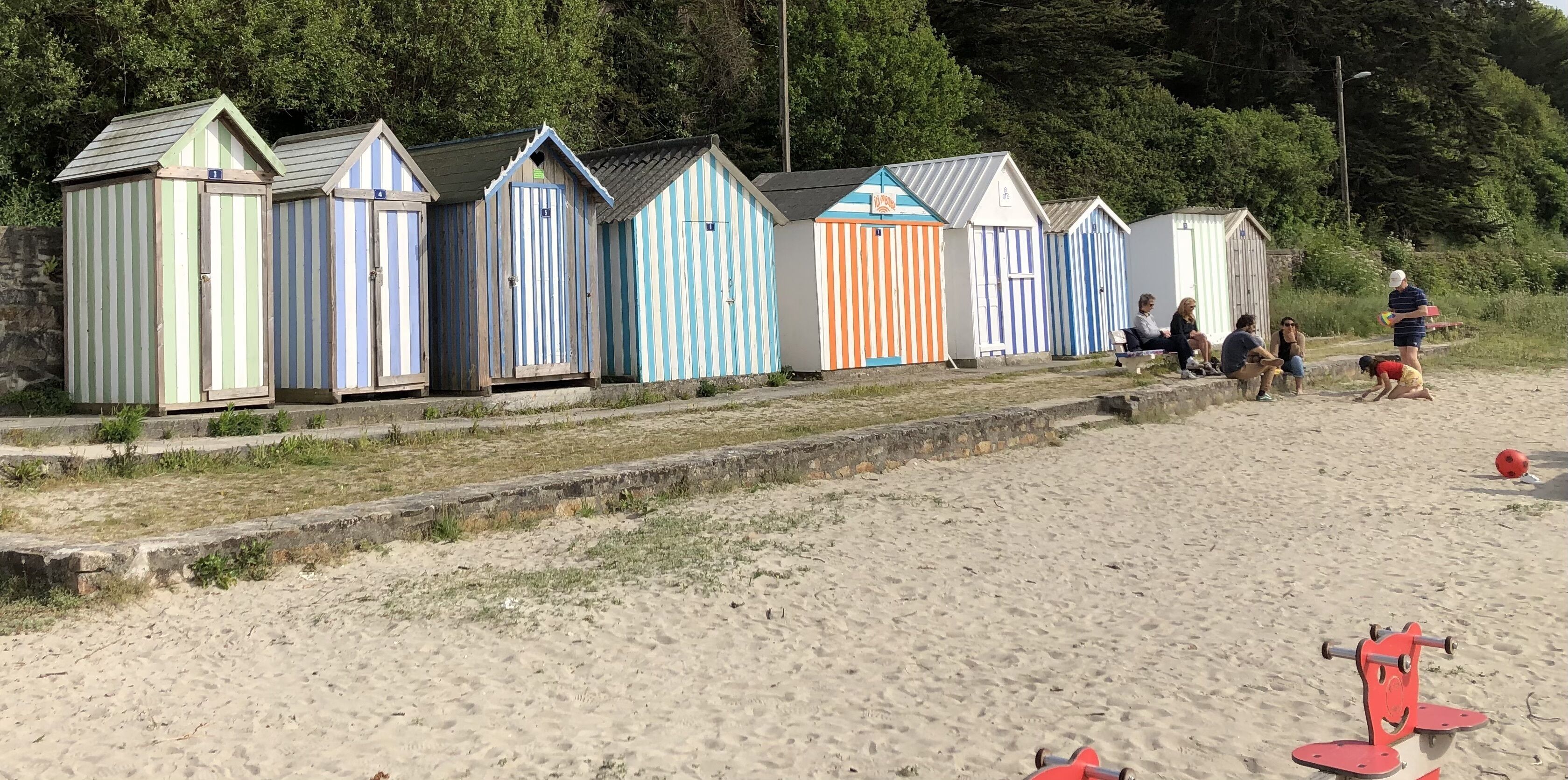 A row of colorful beach huts with a sandy beach and a small water body in the background.