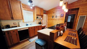 Una cucina compatta con mobili in legno, un lavello e un fornello. Sono presenti luci a sospensione e una finestra con persiane.
