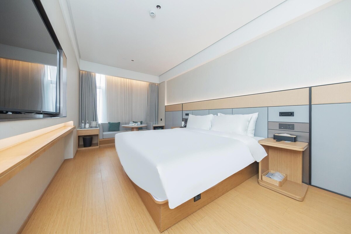 A modern hotel room with a large bed, bedside tables, a flat-screen TV, and a seating area with curtains.