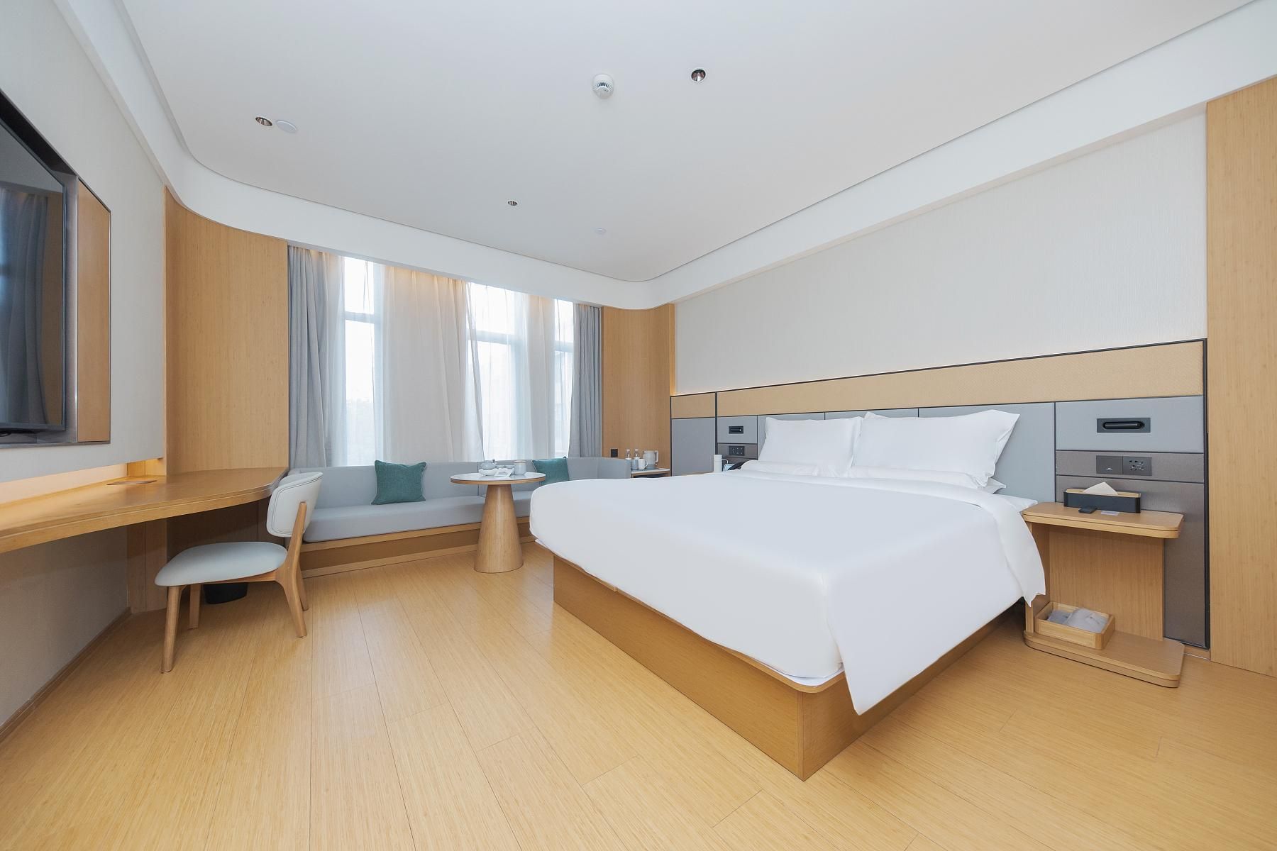 A modern hotel room with a large bed, a desk, a chair, a small table, and a window with curtains.