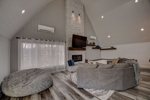 Ein geräumiges Loft mit hohen Decken, einem Kamin aus Stein, einem großen grauen Sofa und einem gemütlichen Sitzsack.