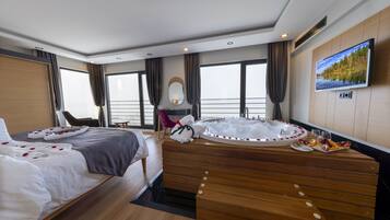 Habitación de hotel moderna con una cama grande, una mesita de madera con hidromasaje, televisor y vistas al mar.