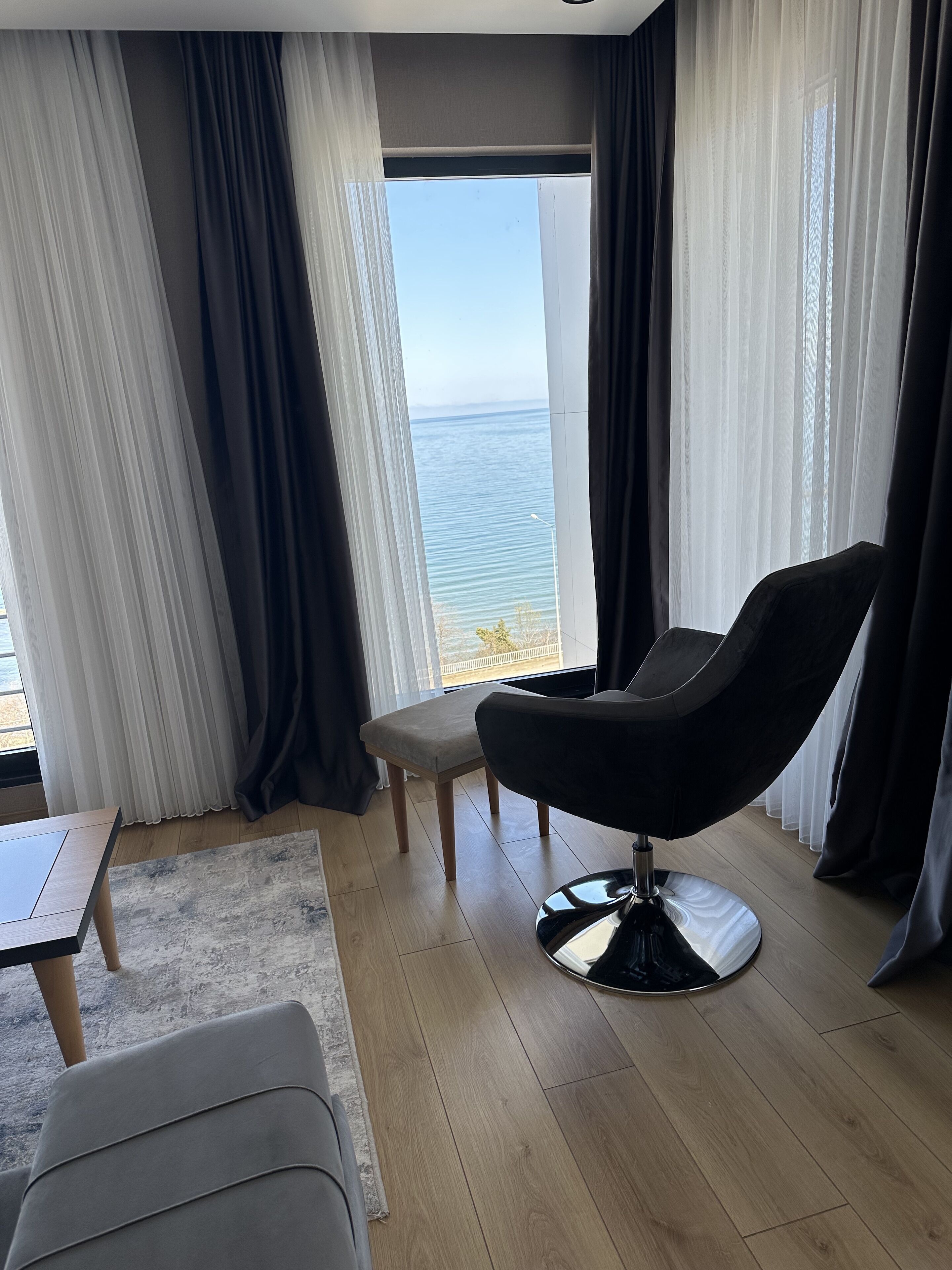 Una habitación moderna con vista al mar, que incluye una silla negra, una mesita pequeña y suelo de madera.