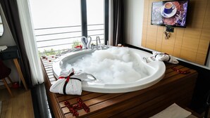 A bathroom with a large bathtub filled with bubbles, a wooden vanity, a TV mounted on the wall, and a view of the outdoors. - Şehrar Suit Hotel (Surmene)