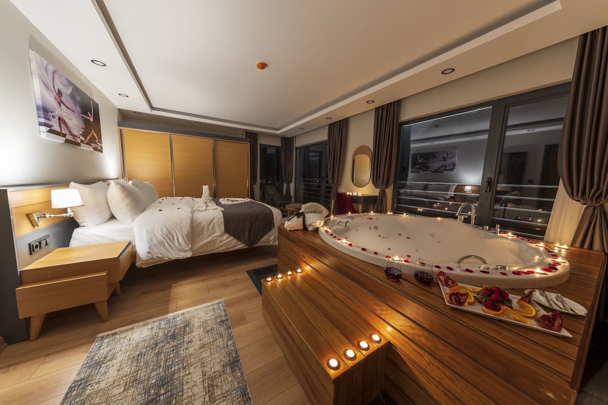 A modern hotel room with a large bathtub, a bed, a bedside table, and a view of the city at night.