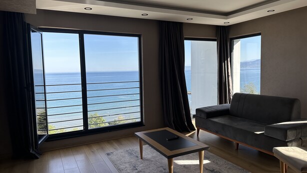 A modern living room with a large window offering a view of the sea, a sofa, a coffee table, and a rug. - Şehrar Suit Hotel (Surmene)