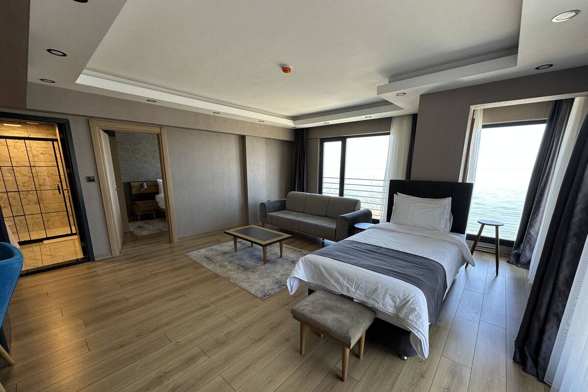 A modern hotel room with a large bed, a sofa, a small table, and a view of the sea.