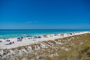 A beach with white sand, clear blue water, and a grassy dune in the foreground.