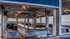 A covered outdoor seating area with wooden benches and tables, a blue awning, and a view of the sea. - Private Beach Access~2 pools~tennis court~Balcony view~Short Walk to Beach (Destin)