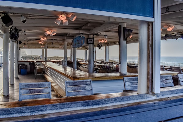 A covered outdoor seating area with wooden benches and tables, a blue awning, and a view of the sea.