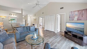 A spacious living room with a grey sofa set, a glass coffee table, a flat-screen TV, and a ceiling fan. - Private Beach Access~2 pools~tennis court~Balcony view~Short Walk to Beach (Destin)