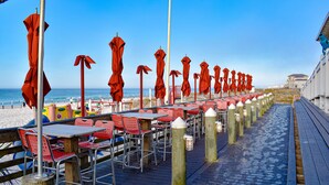 A seaside restaurant with outdoor seating, red umbrellas, and a wooden deck. - Private Beach Access~2 pools~tennis court~Balcony view~Short Walk to Beach (Destin)