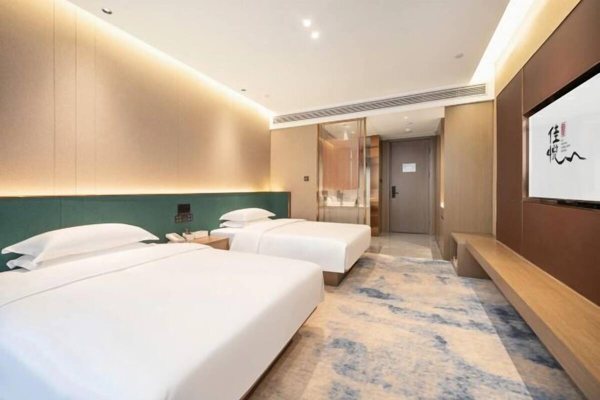 Une chambre d’hôtel avec deux lits, un grand miroir, un banc en bois et un téléviseur fixé au mur.