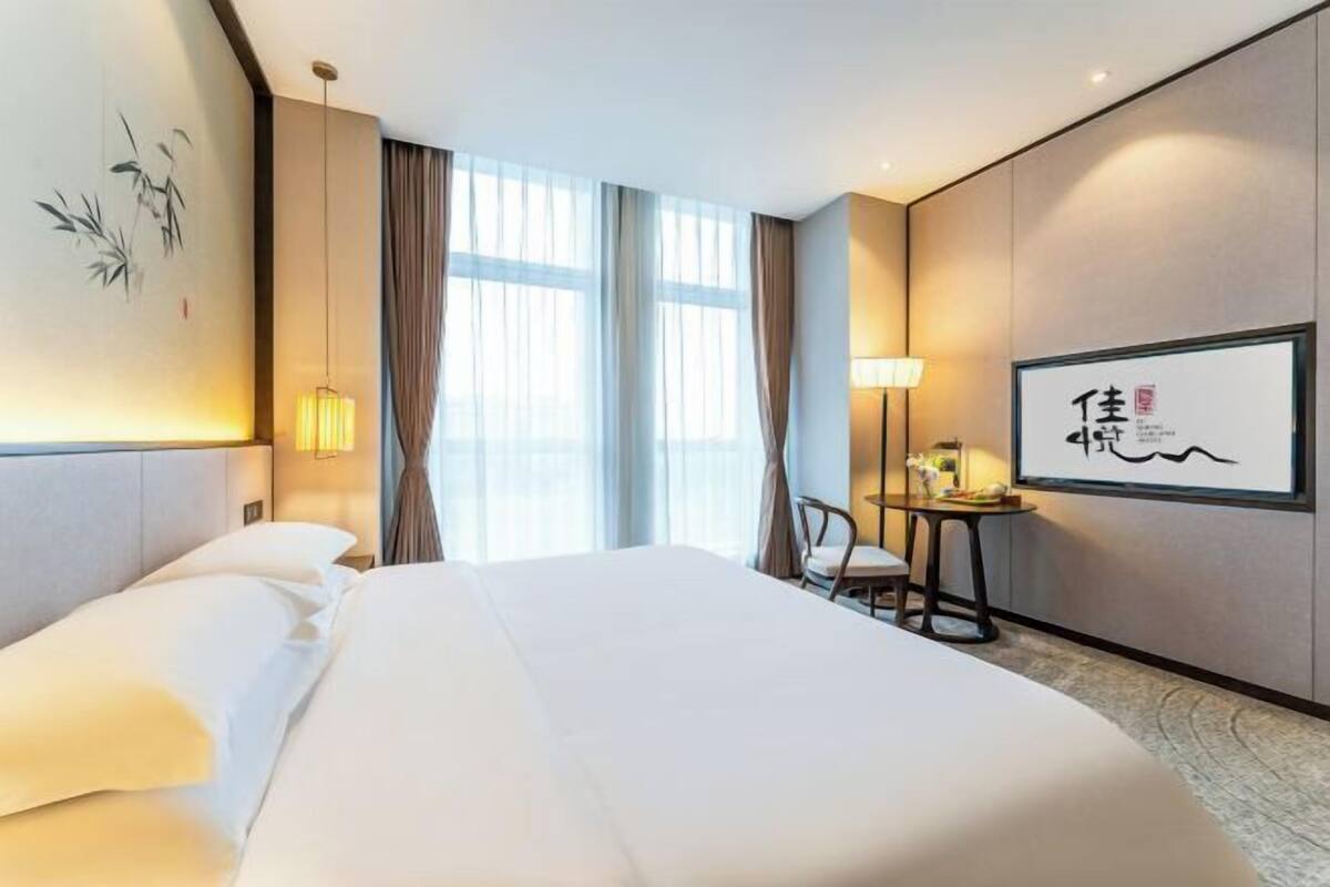 Une chambre d’hôtel avec un grand lit, un bureau avec une chaise, une lampe et un mur décoré d’œuvres calligraphiques.