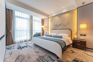Habitación de hotel con una cama grande, una mesita de noche, una lámpara y vistas a la ciudad desde la ventana.