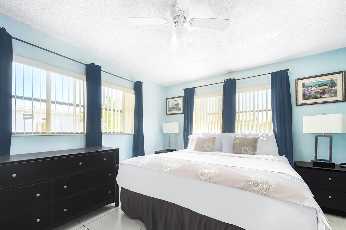 A bedroom with a bed, two bedside tables, a dresser, a ceiling fan, and windows with curtains.