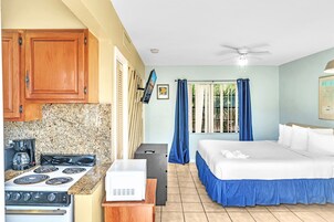 Ett kompakt hotellrum med ett litet kök, en säng, en takfläkt och ett fönster med gardiner.