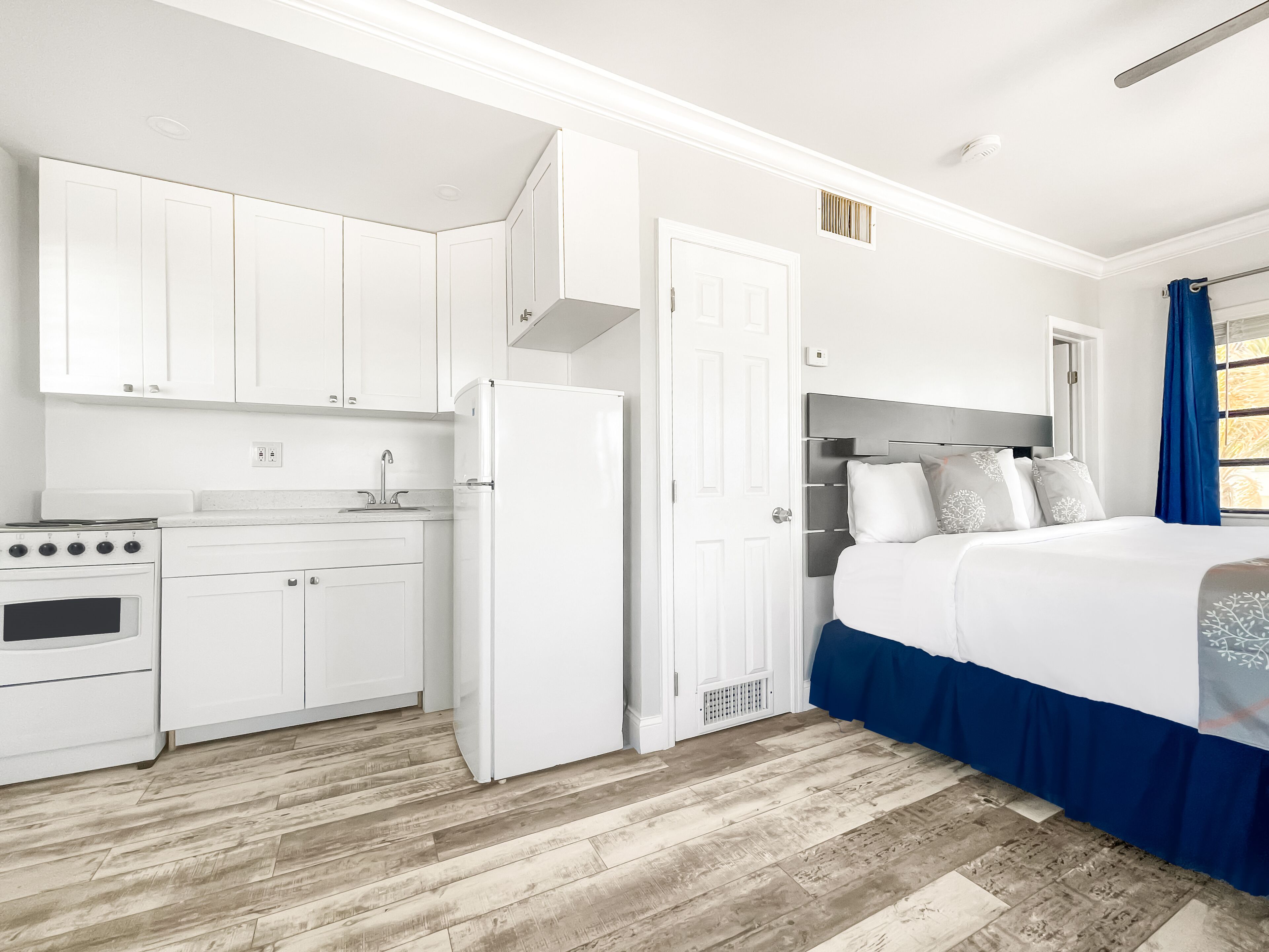 A compact hotel room with a kitchenette, a bed with a blue bed skirt, and a window with blue curtains.
