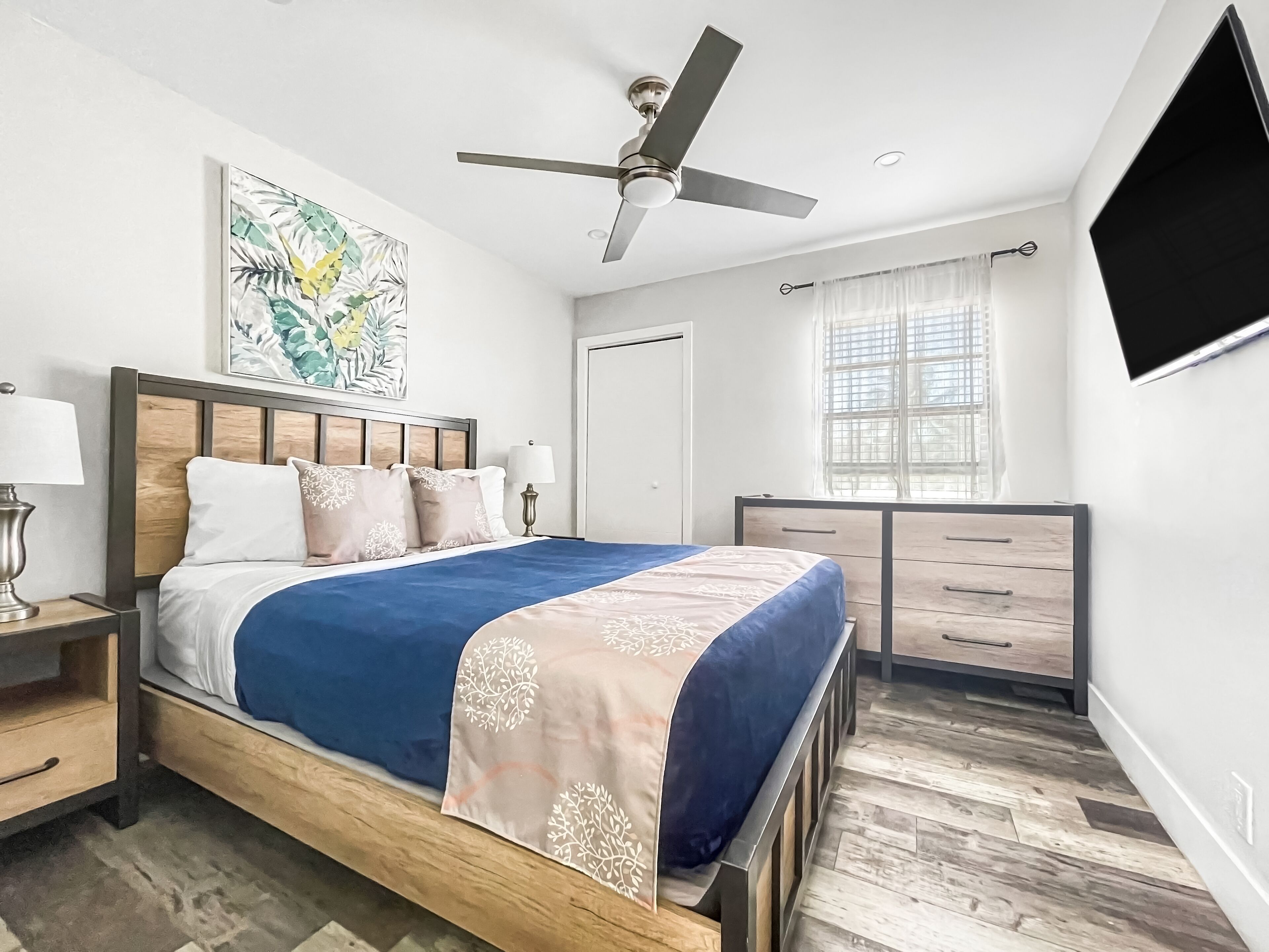 A modern bedroom with a bed, nightstands, a ceiling fan, a window with curtains, and a flat-screen TV.