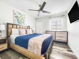 A modern bedroom with a bed, nightstands, a ceiling fan, a window with curtains, and a flat-screen TV.