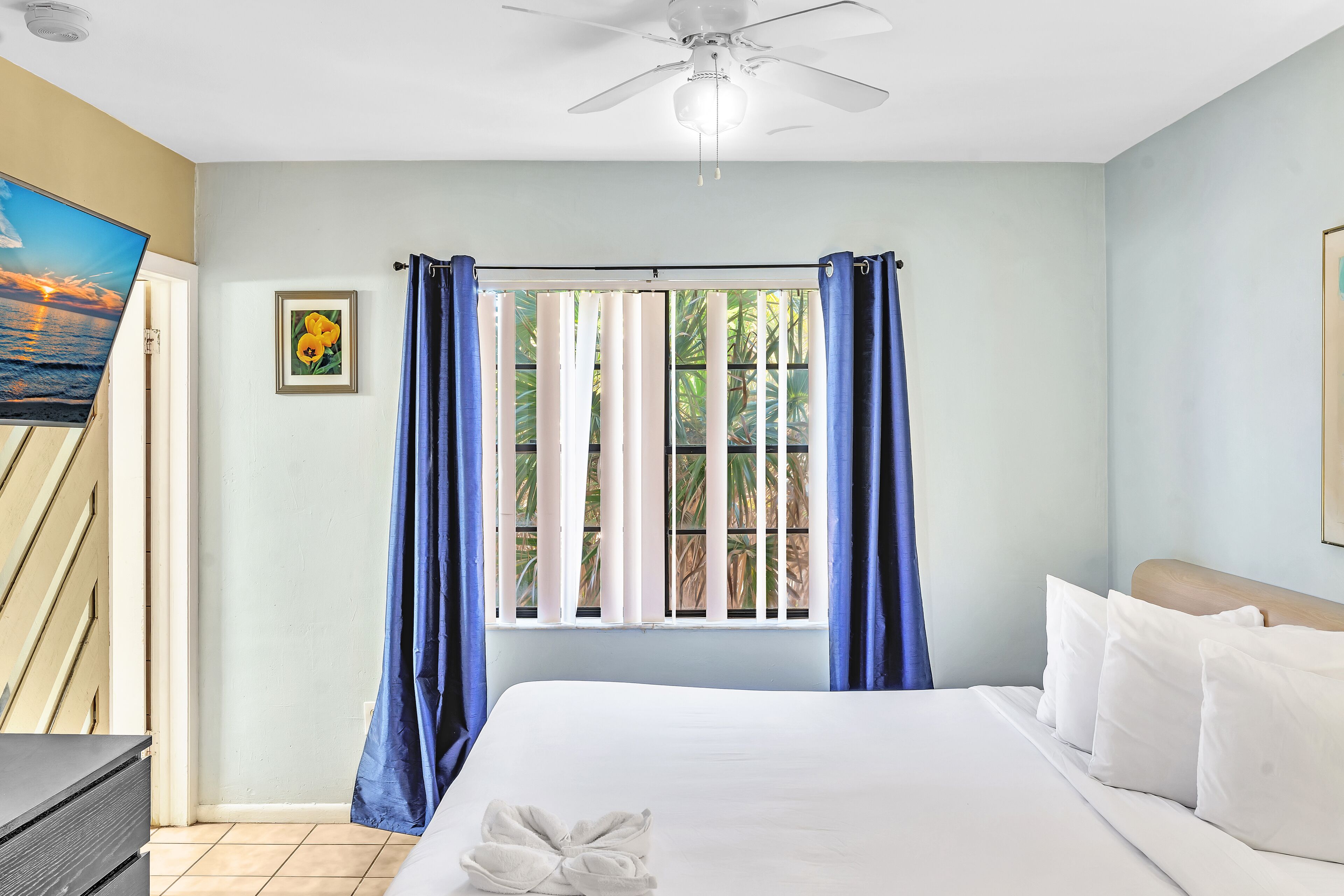 A hotel room with a bed, a ceiling fan, a window with curtains, a painting on the wall, and a door.