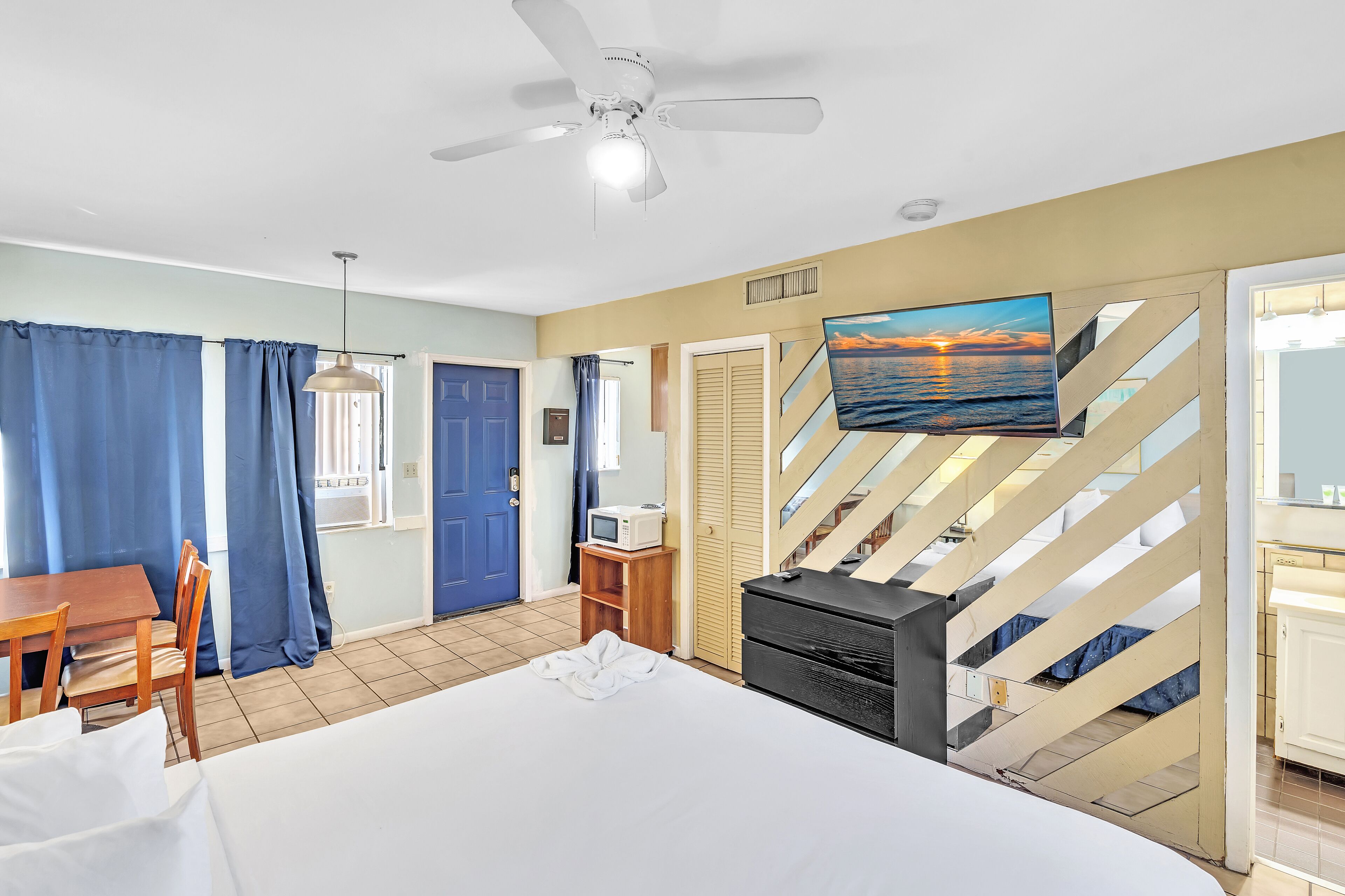 Habitación de hotel con cama, escritorio, televisor, ventilador de techo y vista al mar a través de una ventana.