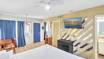 Habitación de hotel con cama, escritorio, televisor, ventilador de techo y vista al mar a través de una ventana.