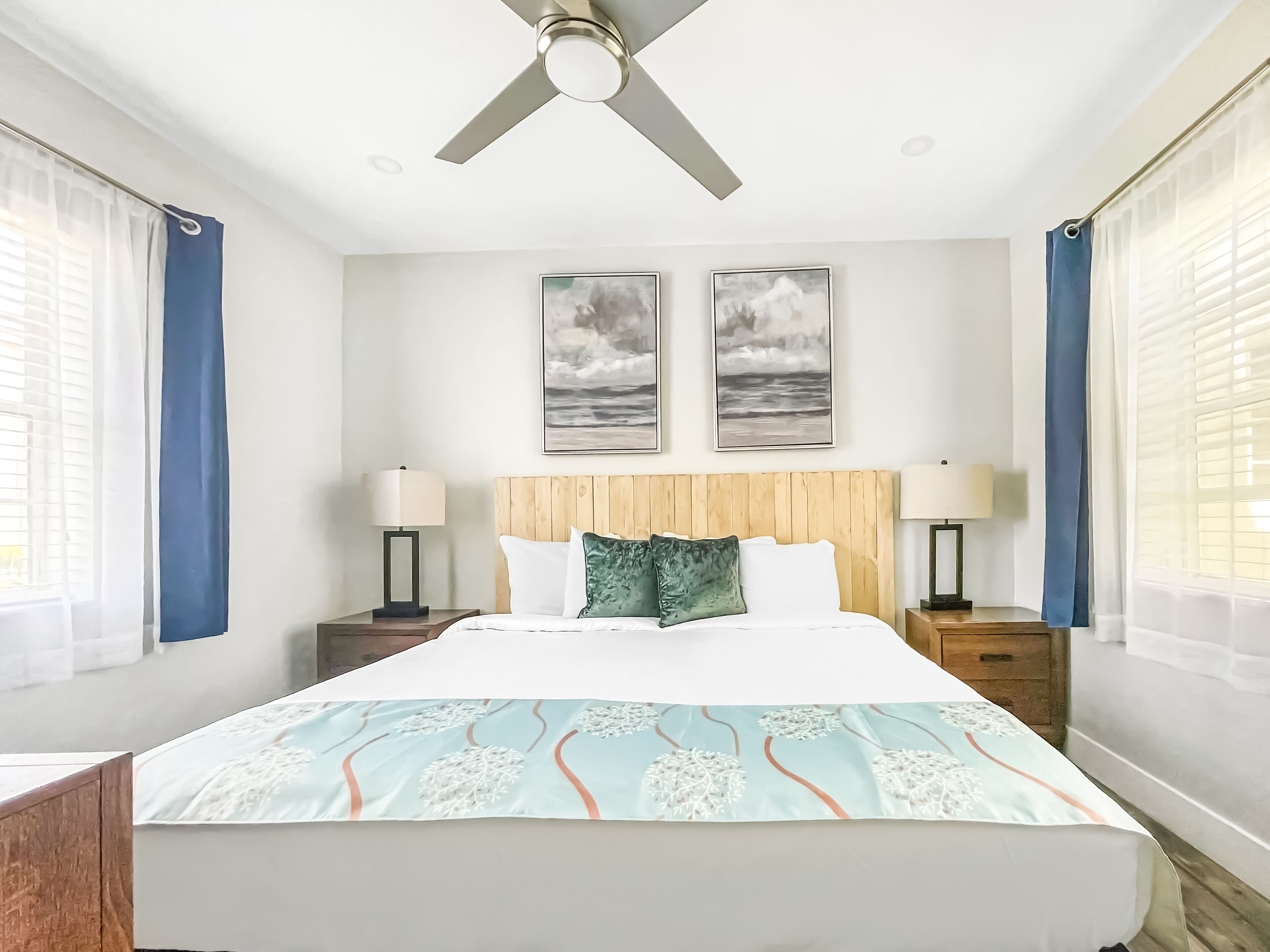A hotel room with a large bed, two wall-mounted paintings, a ceiling fan, and a window with curtains.
