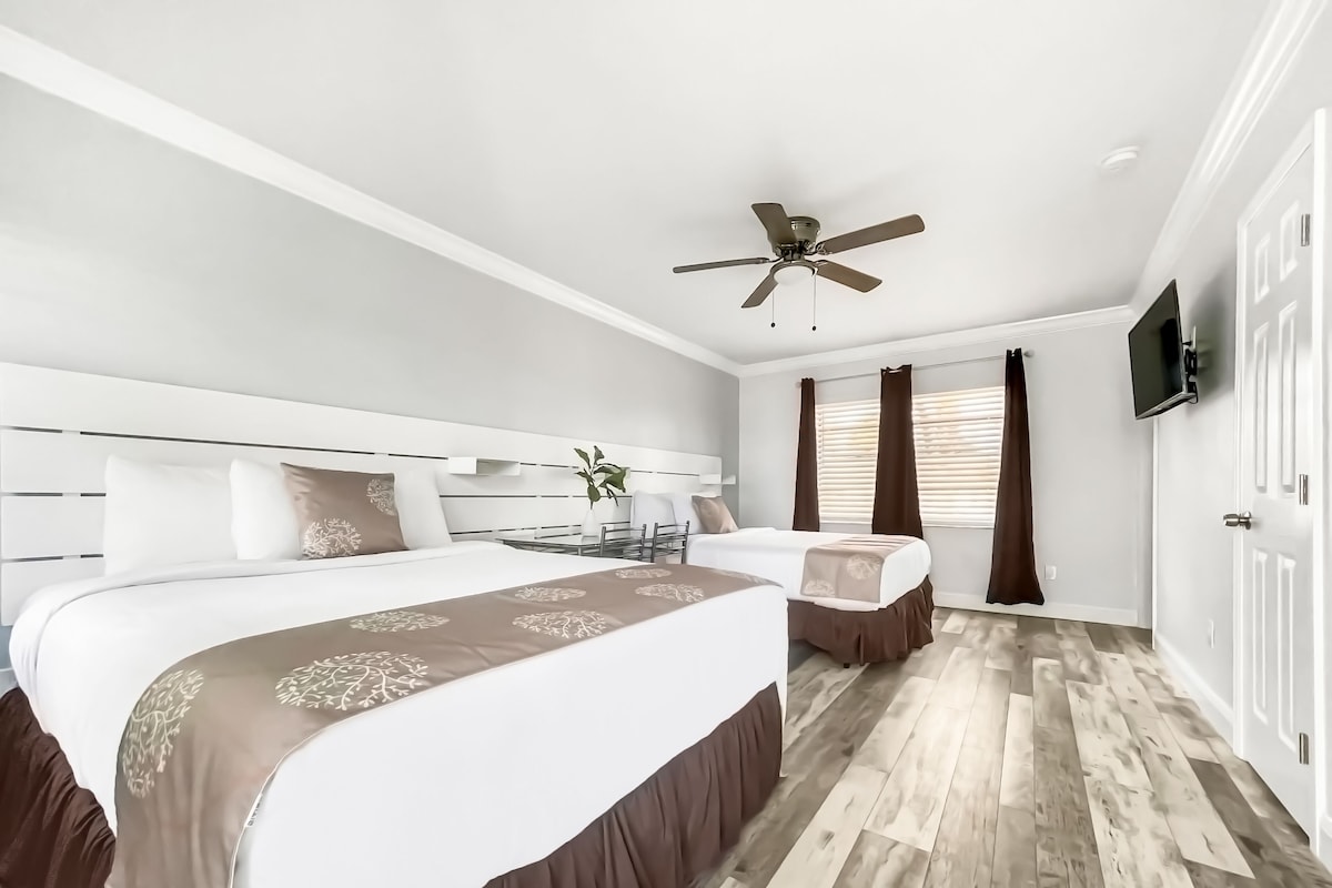 Une chambre d’hôtel avec un grand lit, un lit plus petit, un bureau, une chaise, un ventilateur de plafond, une télévision et une fenêtre avec des rideaux.