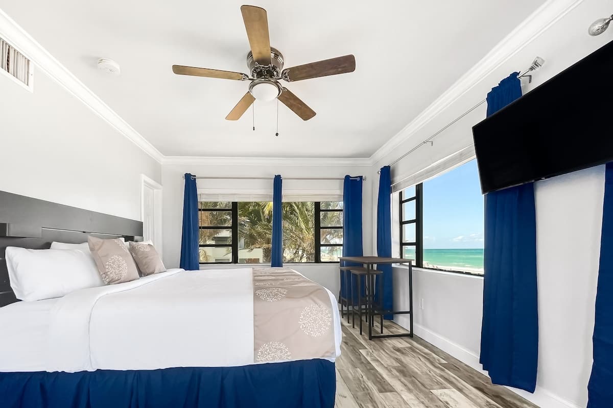 A bedroom with a bed, a ceiling fan, a view of the ocean, and a balcony with a table.