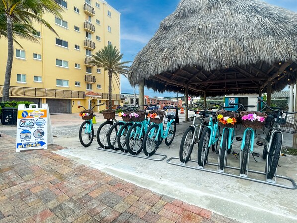 A beachfront hotel with a thatched-roof structure, bicycles with baskets, and a rental sign.