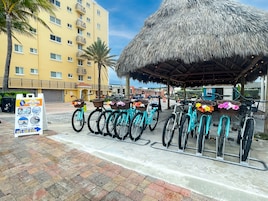 A beachfront hotel with a thatched-roof structure, bicycles with baskets, and a rental sign.