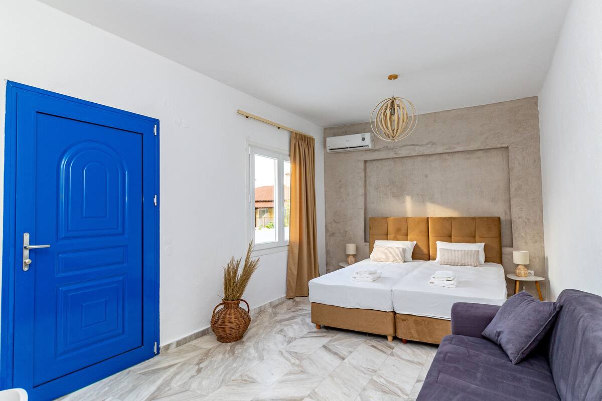 Een slaapkamer met een blauwe deur, een bed met twee kussens, een nachtkastje met een lamp en een potplant.