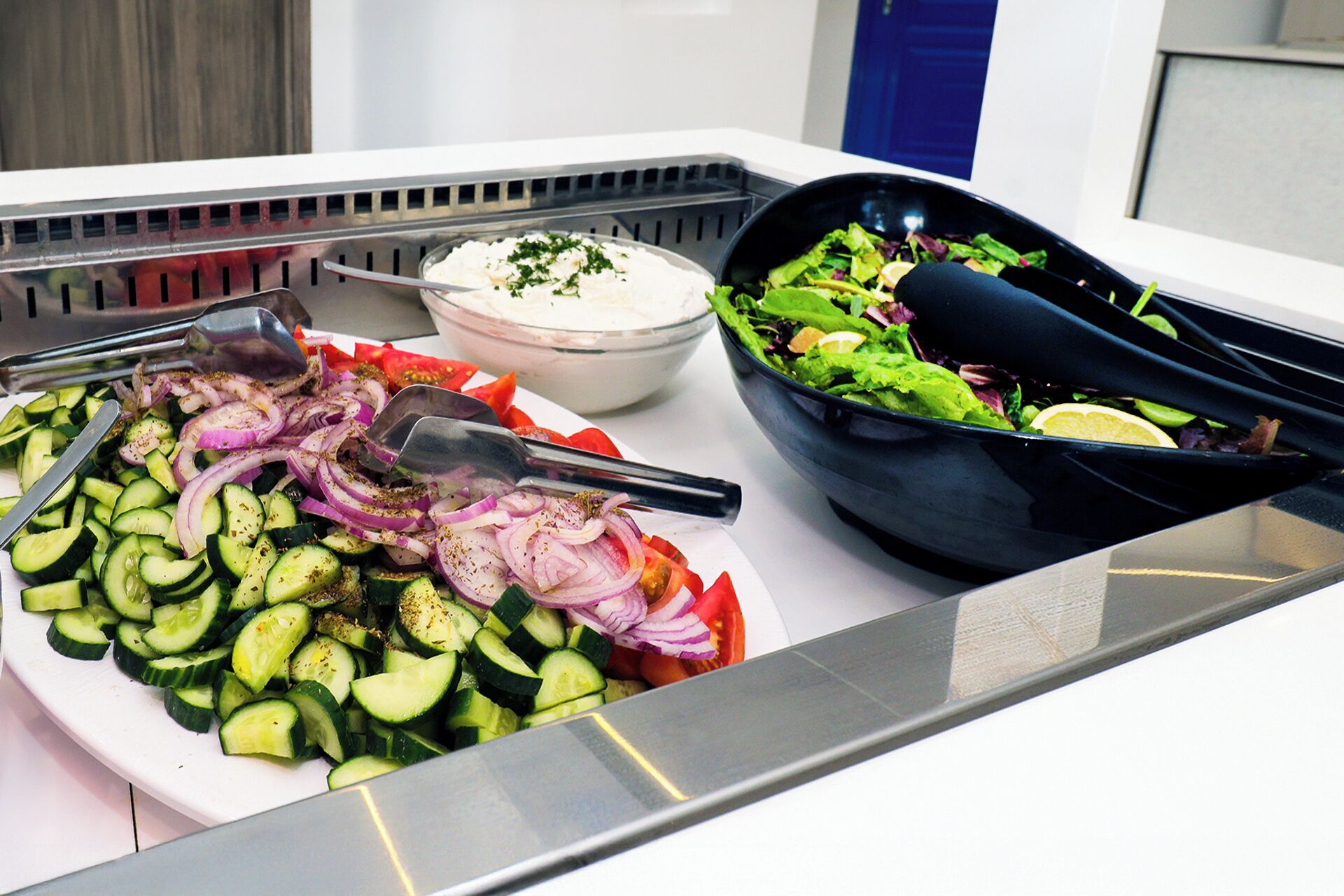 Una mesa de bufé con diversos platos, incluyendo pepinos en rodajas, cebollas rojas y una ensalada en un tazón negro.