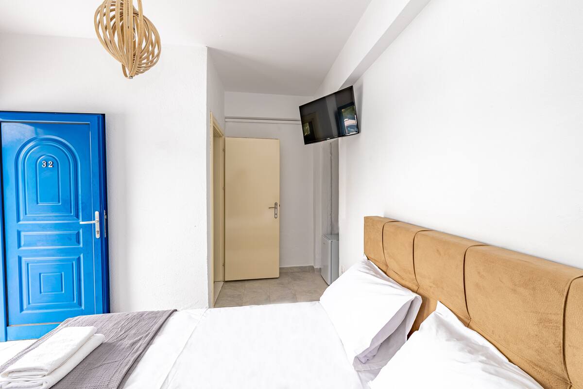Een slaapkamer met een blauwe deur, een bed met witte lakens, een televisie aan de muur en een houten hoofdbord.