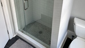 Une douche fermée par des parois de verre, avec un sol carrelé, des toilettes et une petite étagère contenant des articles de toilette.
