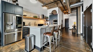Une cuisine moderne dotée d’appareils électroménagers en acier inoxydable, d’un îlot central avec des tabourets de bar et de conduits apparents au plafond.