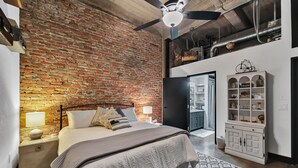 Une chambre avec un mur en briques, un lit avec une tête de lit, un ventilateur de plafond, une table de chevet, une lampe, une armoire et un tapis à motifs.