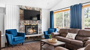 Ein Wohnzimmer mit Kamin aus Stein, blauen Sesseln, einem braunen Ledersofa und einem großen Fenster mit Vorhängen.