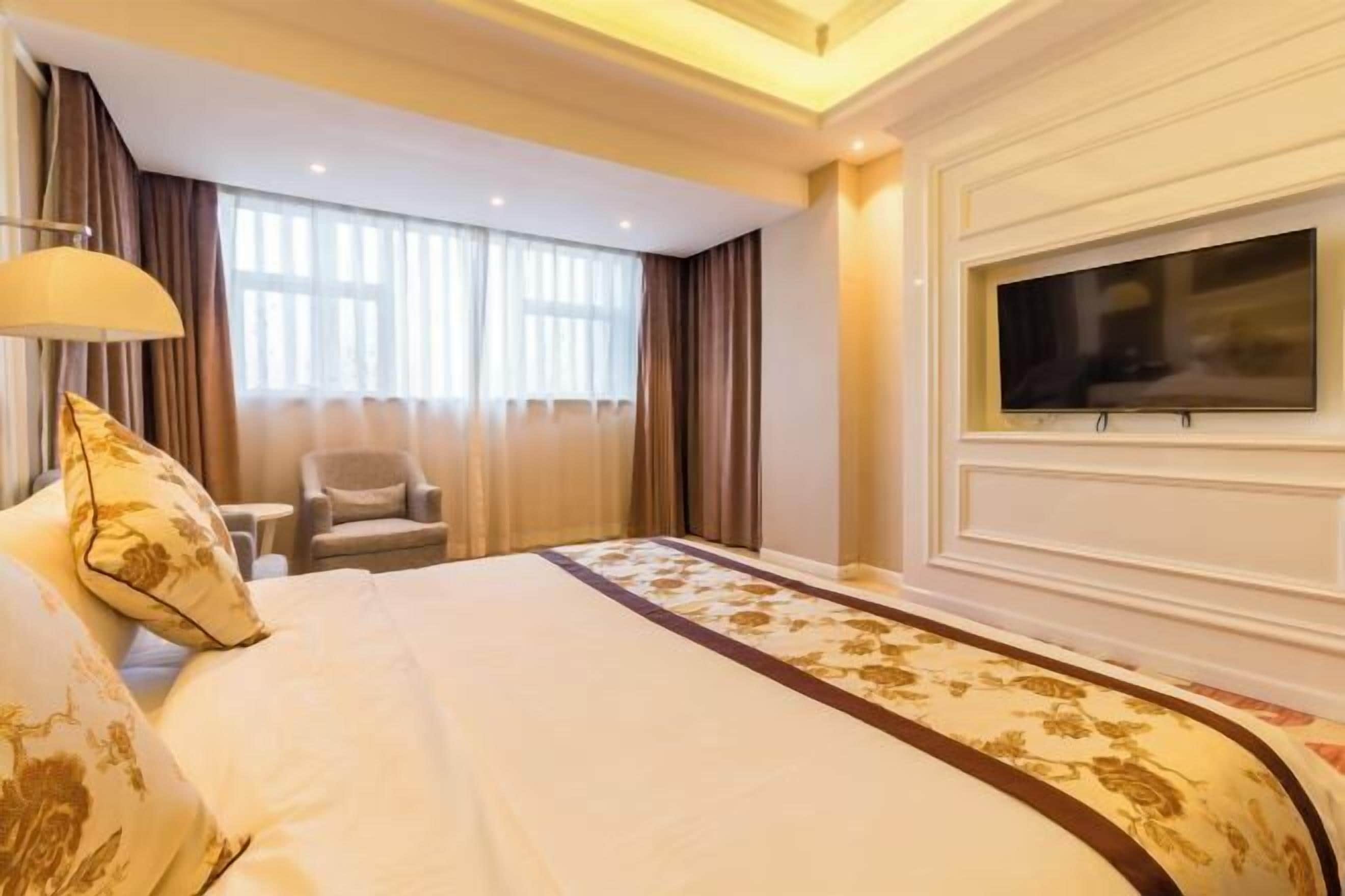 A hotel room with a bed, a TV, a chair, and a window with curtains.