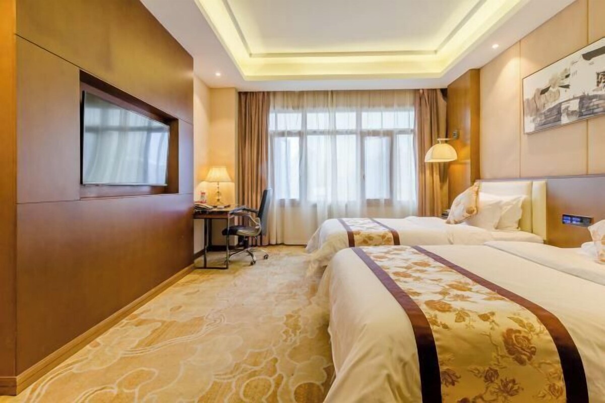 A hotel room with two beds, a desk, a chair, a TV, a window with curtains, and a lamp.