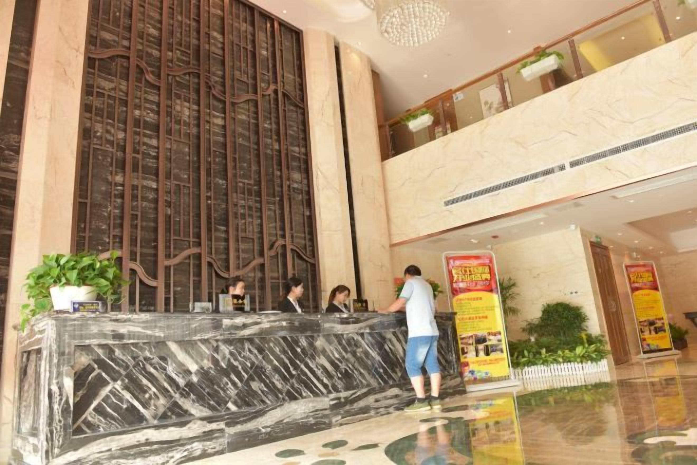 A hotel reception area with a marble counter, a large vertical wooden panel, and a balcony with plants.