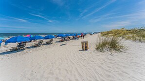 Ein Strand mit blauen Sonnenschirmen und Liegestühlen, einem hölzernen Rettungsschwimmerstand und Dünen im Hintergrund.