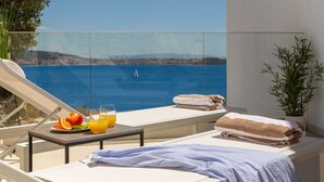 En balkon med frit vue over havet, en liggestol, håndklæder, et bord med frugt og drikkevarer, samt en potteplante.